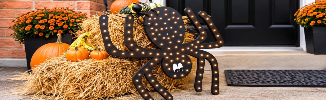 Black Halloween spider decoration illuminated with warm white lights, displayed on hay bales alongside pumpkins and orange fall flowers on a decorated front porch.